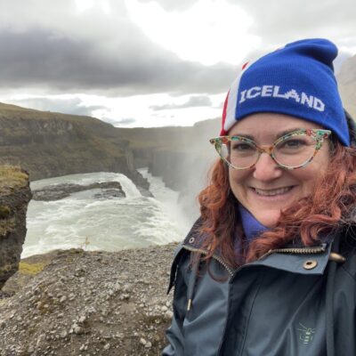 red head curly haired woman wearing glasses and a blue Iceland beanie in front of waterfall